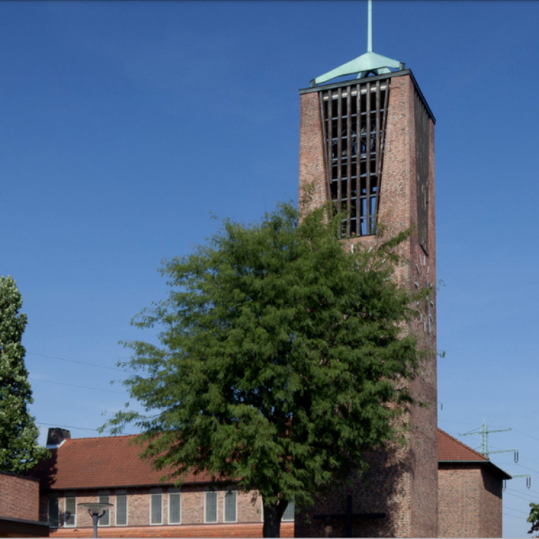 Roter Backsteinturm mit grünem Dach unter blauem Himmel und Bäumen im Vordergrund.