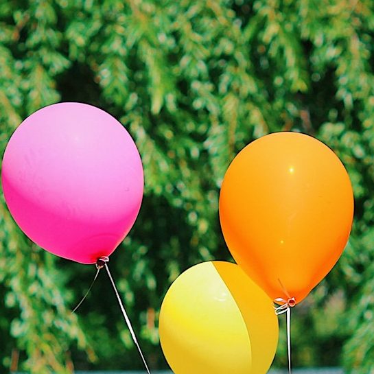 Drei Luftballons in Pink, Orange und Gelb vor grünem Hintergrund.