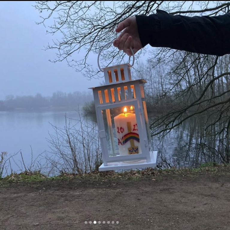 Eine Hand hält eine Laterne mit einer brennenden Kerze am Ufer eines ruhigen Gewässers.