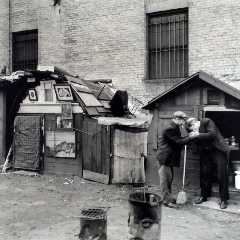 Zwei Männer stehen vor einfachen, improvisierten Unterkünften aus Holz und Material.