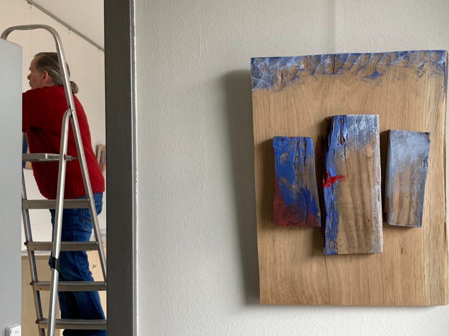 Ein Künstler steht auf einer Leiter, neben einem Holzrelief mit blauen und roten Akzenten.