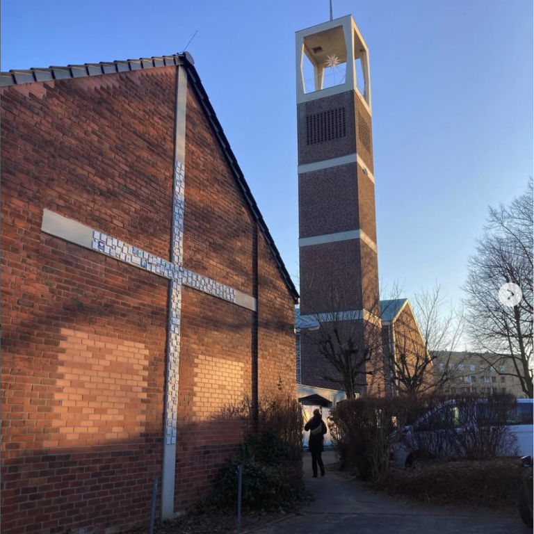 Kirche mit hohem Turm und Kreuz an der Fassade, umgeben von Bäumen und einem klaren Himmel.