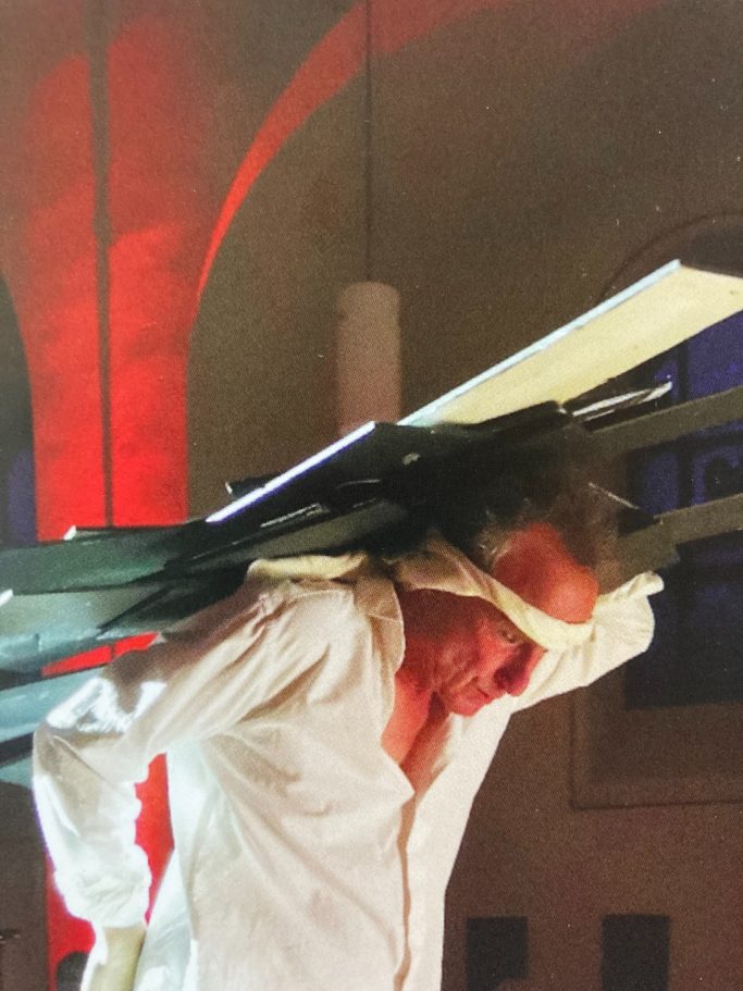 Ein Mann mit einem hölzernen Gestell auf dem Rücken, in einem beleuchteten Raum.