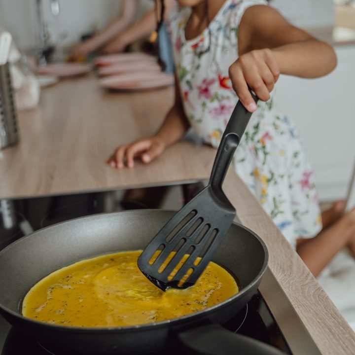 Ein Kind mit blumenbedrucktem Kleid, das mit einem Spatel Eier in einer Pfanne rührt.