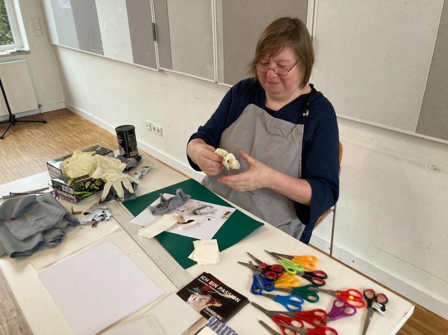 Frau schneidet Papier und arbeitet an einem Bastelprojekt am Tisch. Scheren und Materialien liegen bereit.