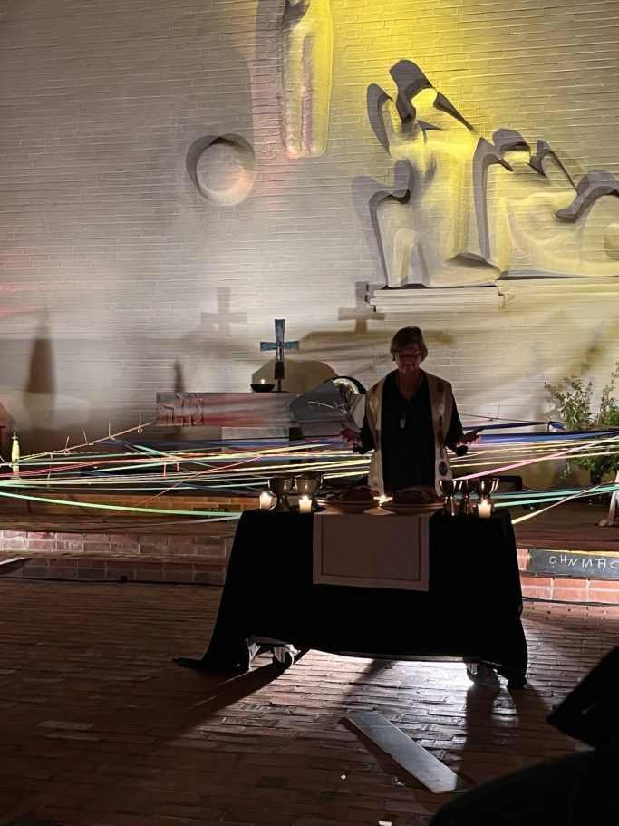 Person steht hinter einem Tisch mit Kerzen, umgeben von bunten Bändern und einer Wand mit Skulpturen.