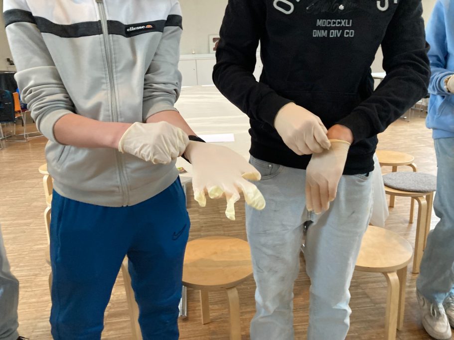 Zwei Personen ziehen Handschuhe an, während sie vor mehreren Holzstühlen stehen.